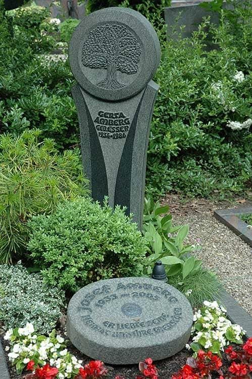 Ein Grabstein mit einem eingemeißelten Baum steht auf einem von Blumen umgebenen Friedhof.