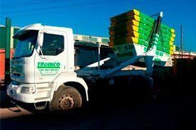 Un camión volquete blanco con contenedores verdes y amarillos en la parte trasera está estacionado al costado de la carretera.