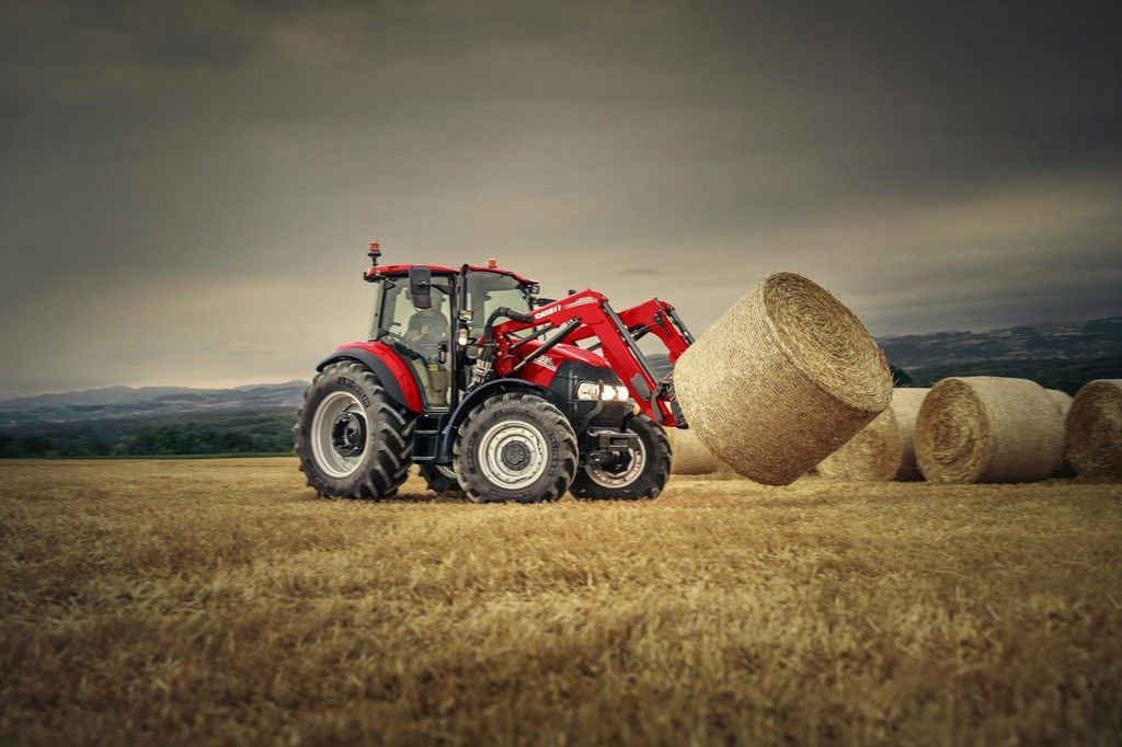 Ein blauer Traktor zieht rote landwirtschaftliche Geräte über ein goldenes Feld unter einem bewölkten Himmel