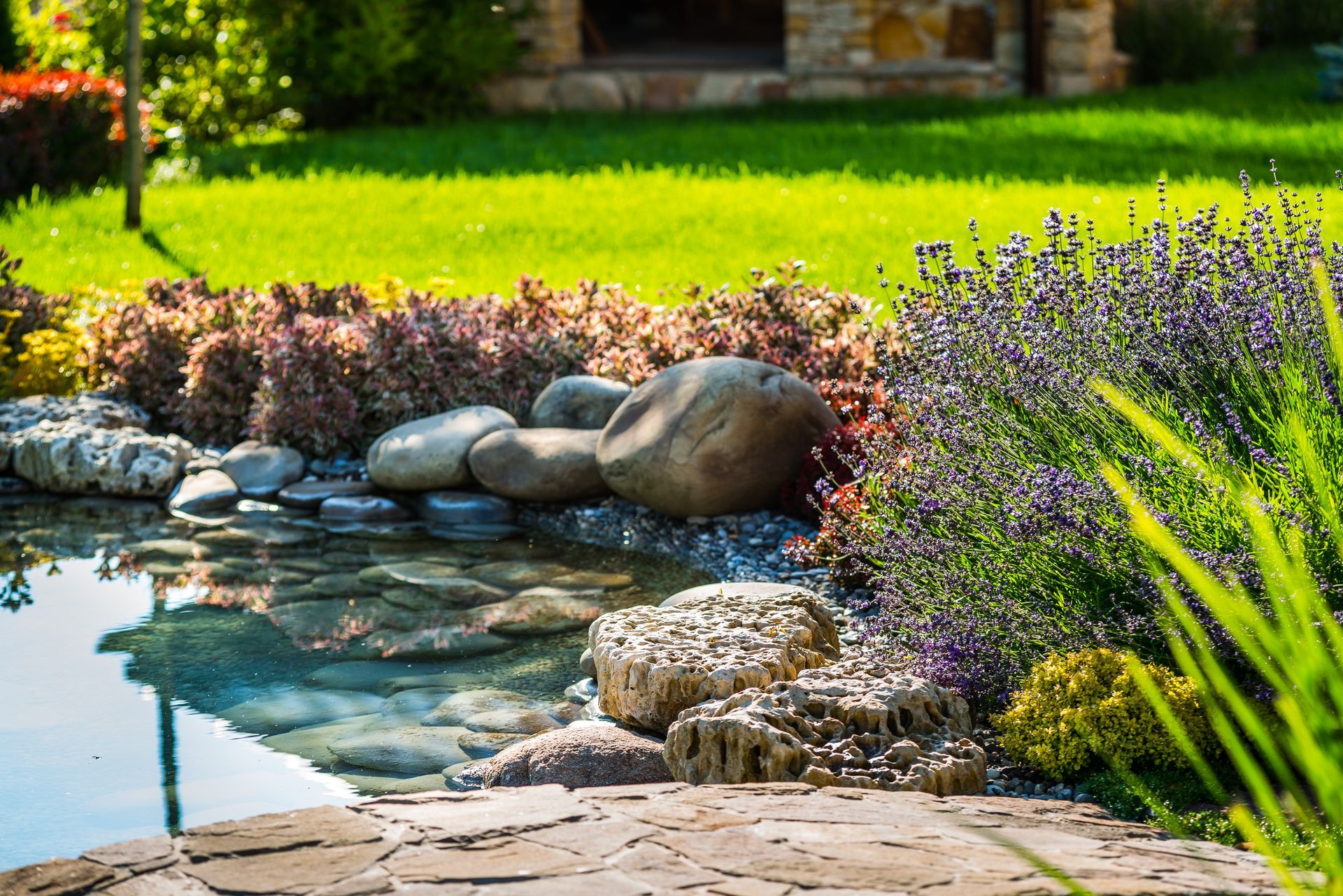 Gartenteich mit Steinen, lila Blumen und grünem Gras neben einer Steinterrasse