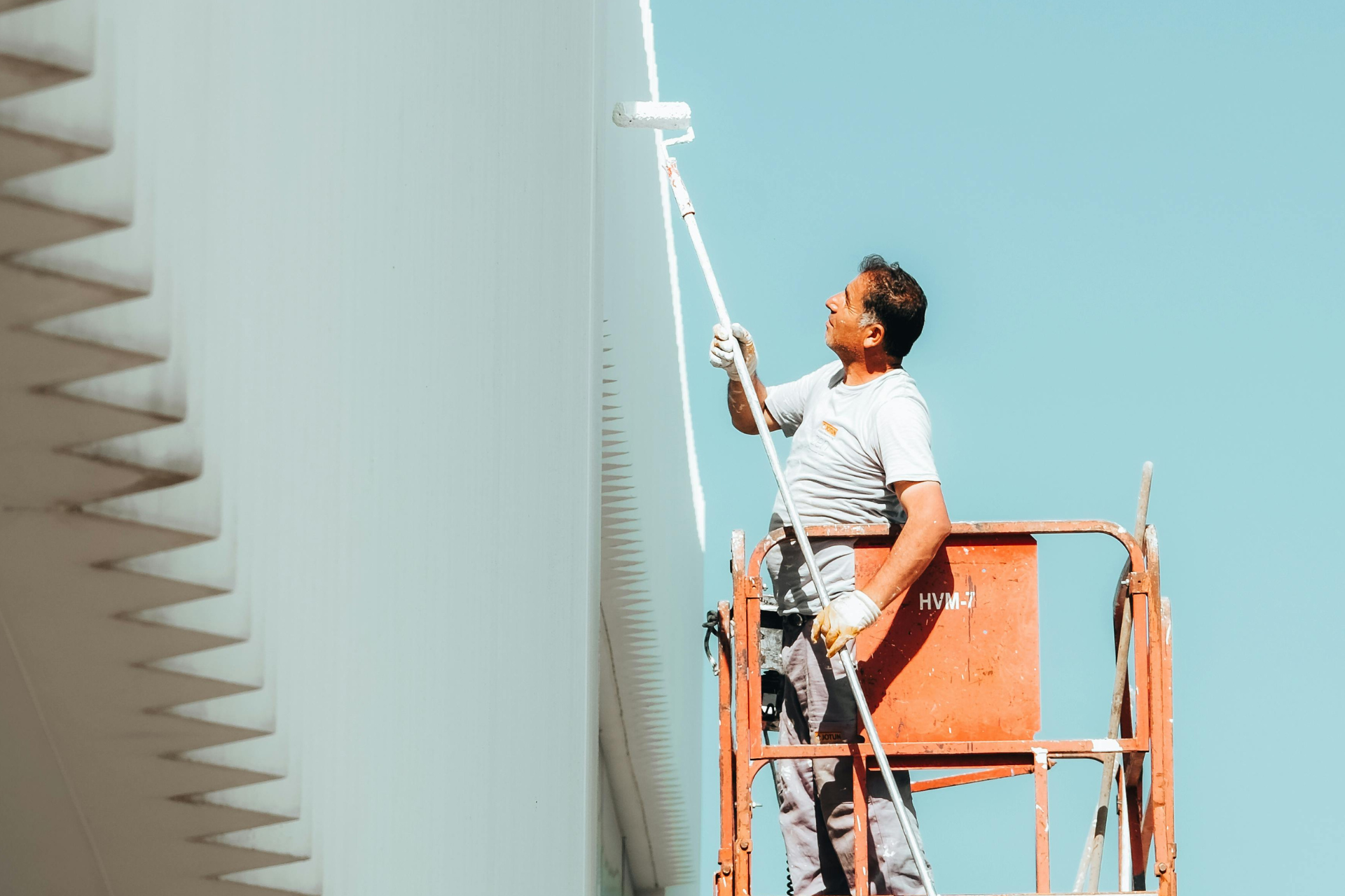 Hombre en un ascensor pintando una estructura alta y blanca con un rodillo contra un cielo azul claro.