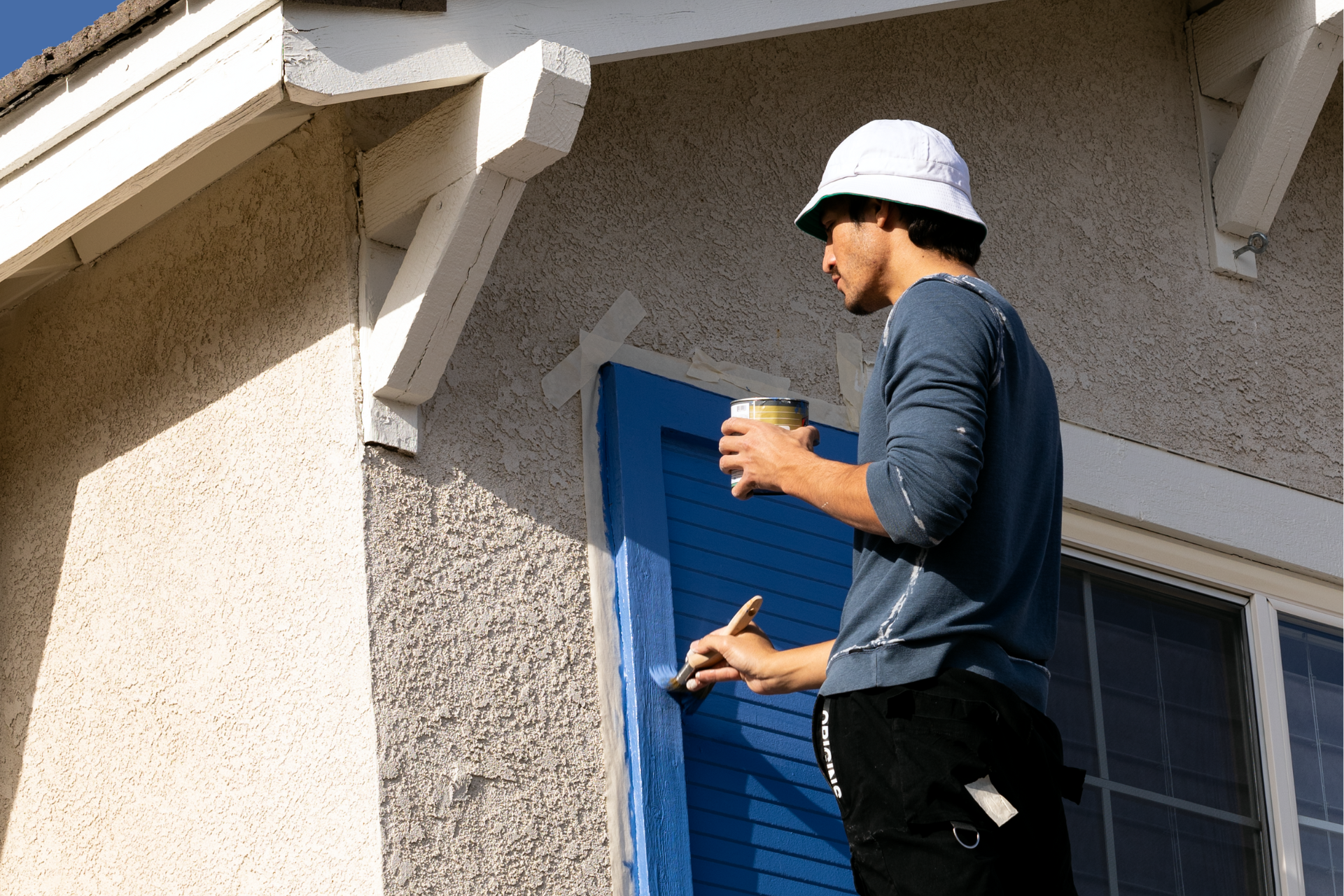 Hombre con casco pintando una puerta azul, con una taza de café en la mano. Fotografía exterior con revestimiento de estuco.