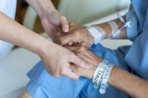 Une personne en blouse d'hôpital bleue, portant un bracelet Risque de chute, tient la main d'un soignant pour se soutenir.