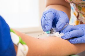 Une personne portant des gants bleus effectue une ponction veineuse, en insérant une aiguille dans un bras avec un garrot vert.