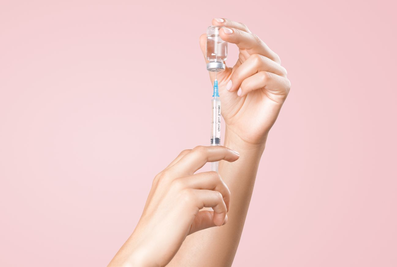Des mains préparent une injection en aspirant un liquide d'une petite fiole en verre sur un fond rose uni.