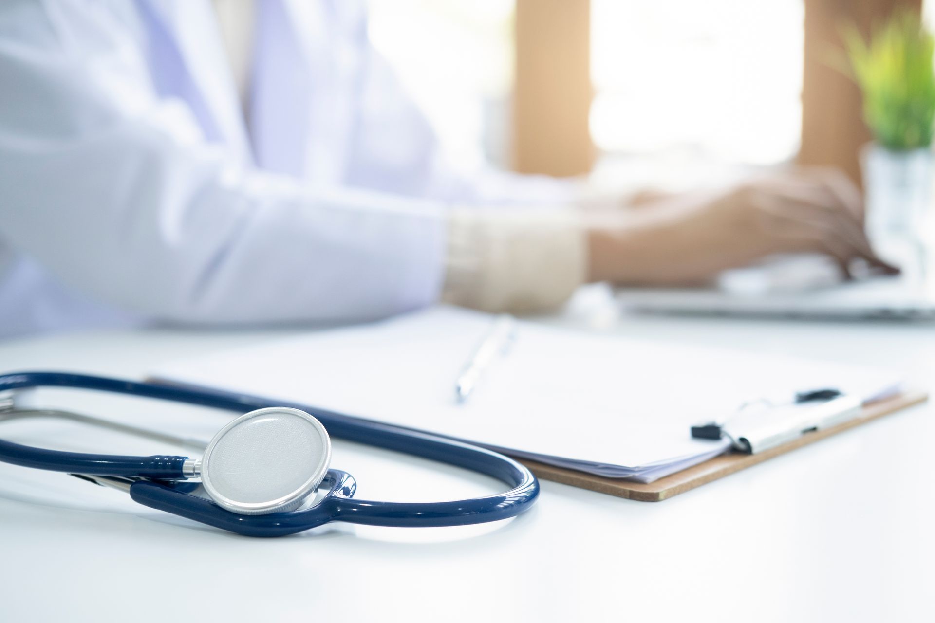 Un stéthoscope est posé sur un bureau au premier plan, tandis qu'un professionnel de la santé travaille sur un ordinateur portable à l'arrière-plan.