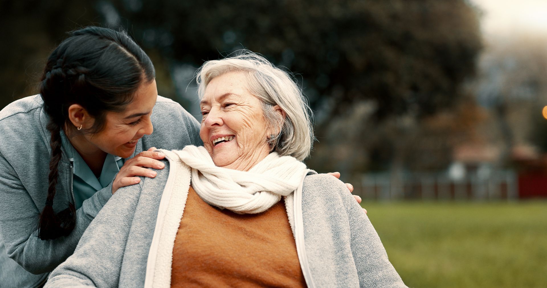 À l'extérieur, une personne souriante aux cheveux foncés pose la main sur l'épaule d'une personne senior souriante portant une écharpe blanche.