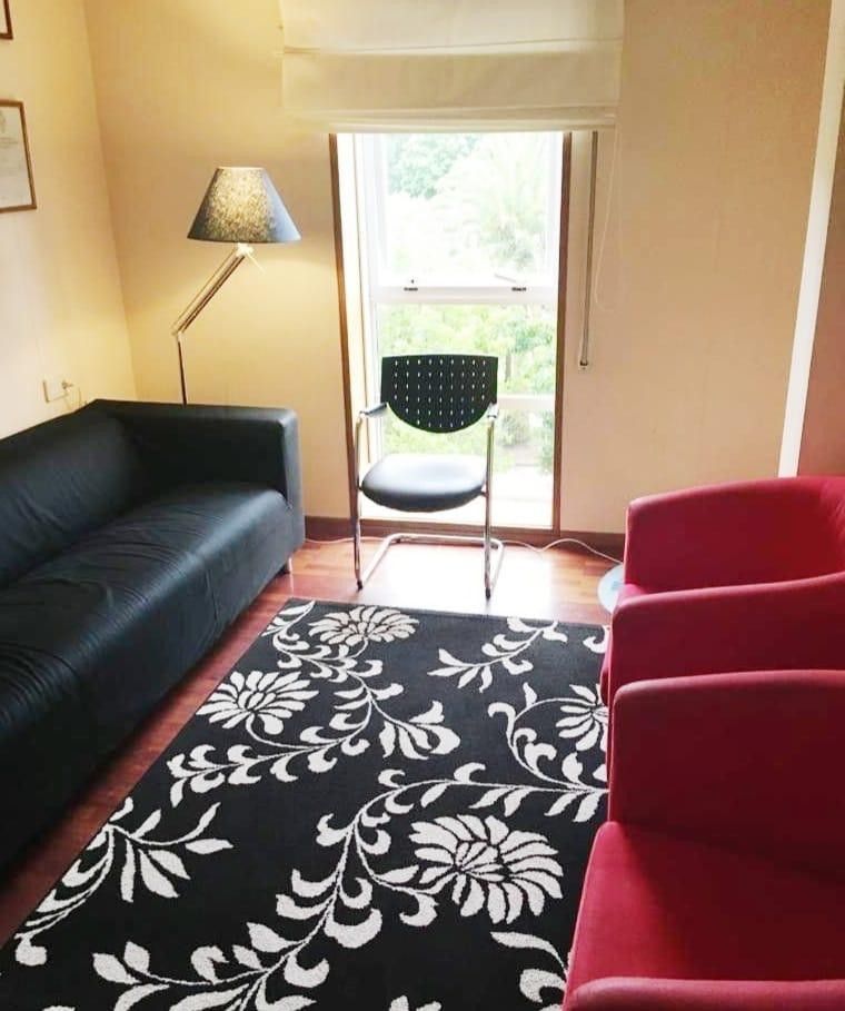 Sala de estar con sofá negro, sillas rojas, alfombra estampada, lámpara de pie y silla junto a una ventana luminosa.