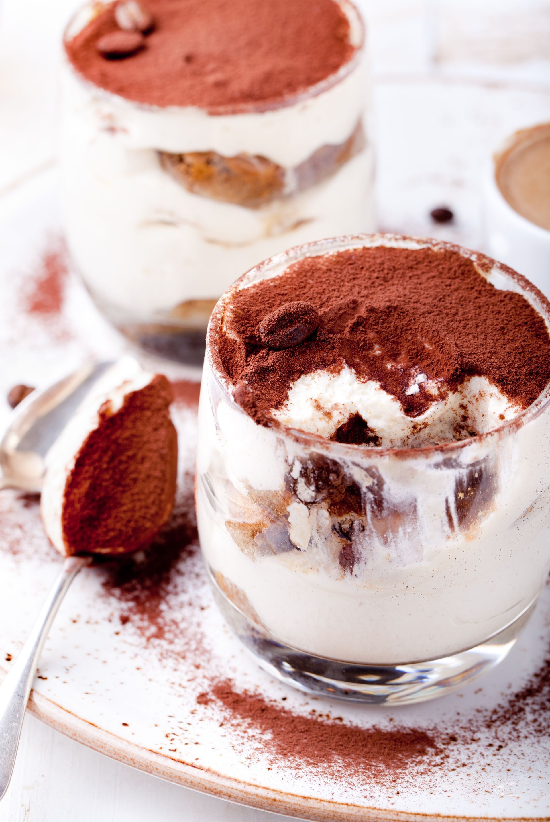 Tiramisu-Desserts in klaren Gläsern, mit Kakaopulver bestreut, mit einem Löffel auf einem Teller.