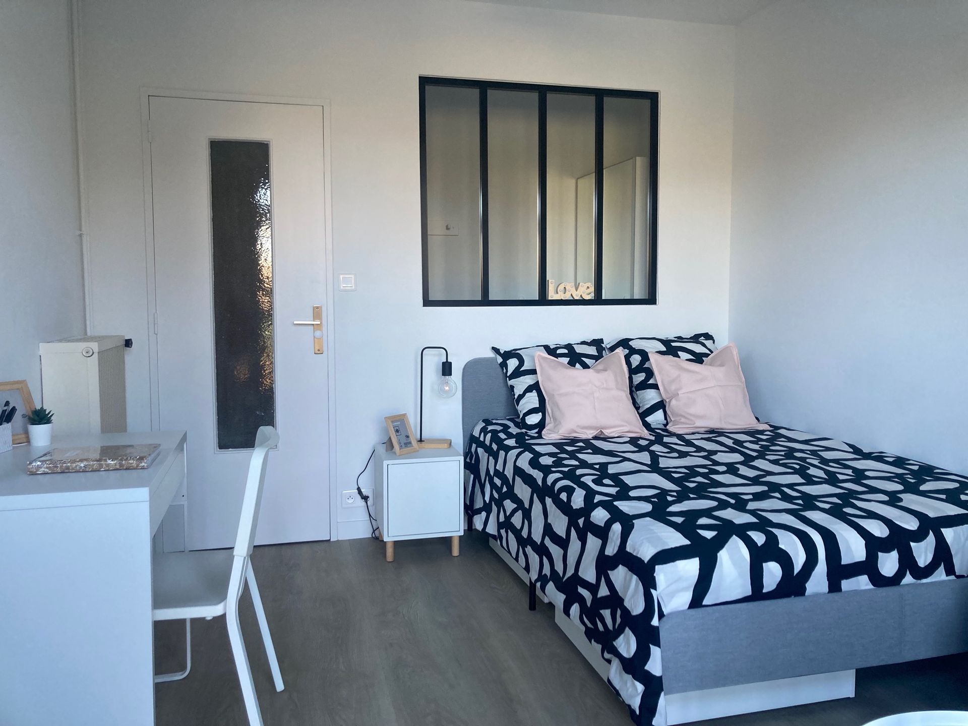 Chambre avec un lit, un bureau et une porte. Couvre-lit à motifs noirs et blancs, murs blancs et une fenêtre.