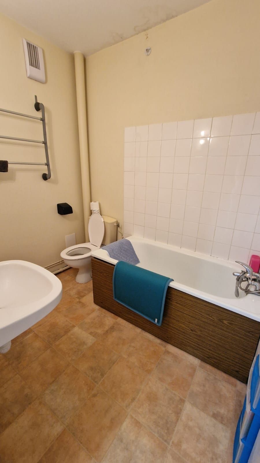 Salle de bains avec baignoire, toilettes, lavabo et porte-serviettes. Murs beiges, sol marron et carrelage blanc.