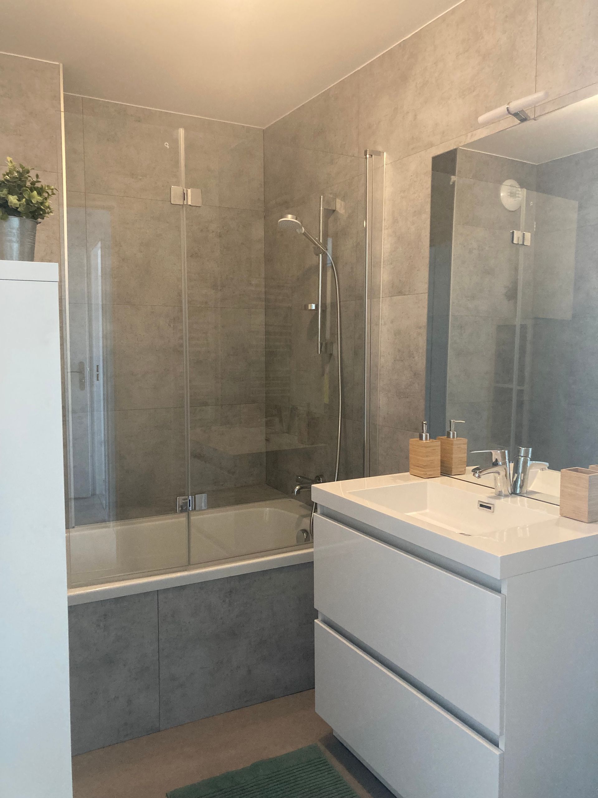 Salle de bains avec murs carrelés gris, meuble-lavabo blanc, baignoire et porte de douche en verre.