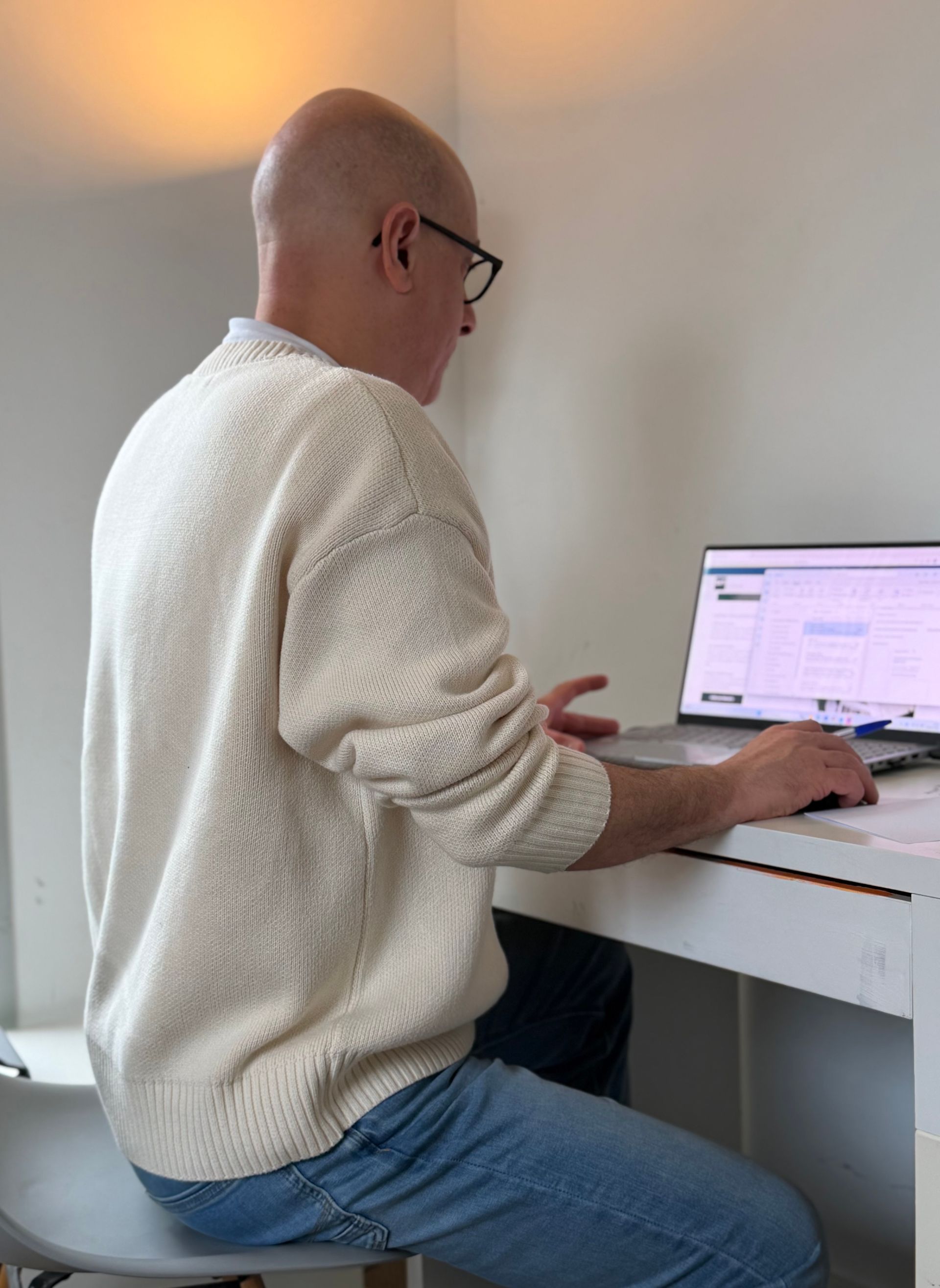 Homme est assis à son bureau, les yeux sur l'écran de son ordinateur portable. Il porte des lunettes, un pull et un jean.