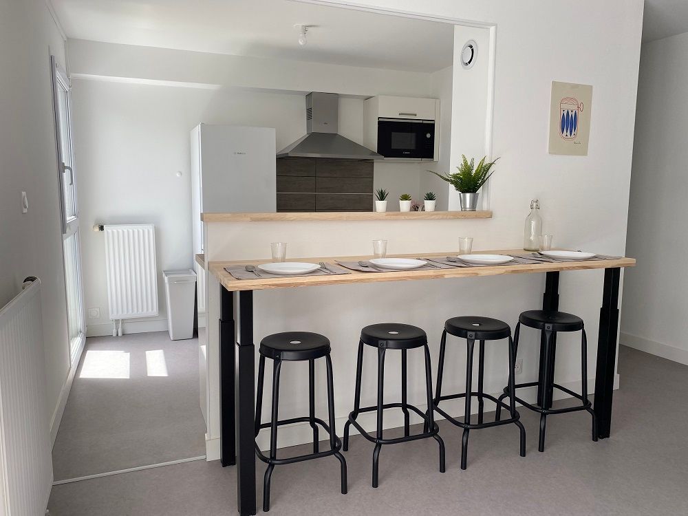 Une cuisine avec un bar, des tabourets et des appareils électroménagers. Murs blancs, sol gris 