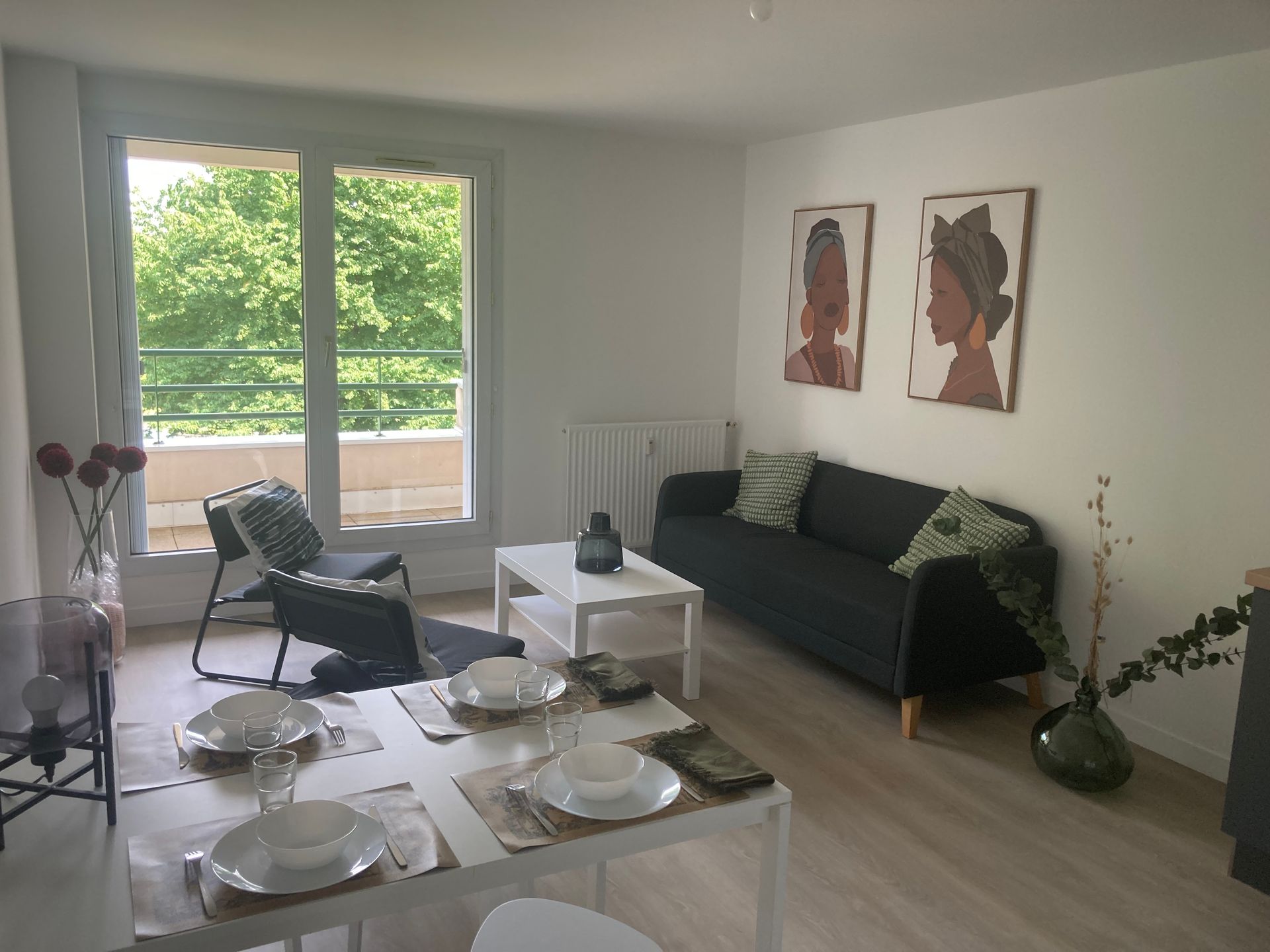 Salon avec canapé noir, table blanche, œuvres d'art et balcon avec végétation.