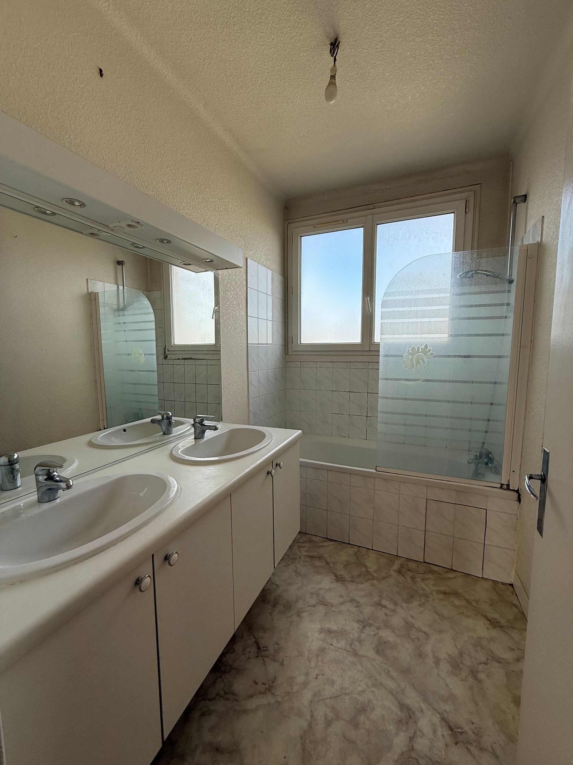Salle de bains avec double vasque, baignoire et fenêtre. Meubles blancs, sol imitation marbre et verre dépoli.
