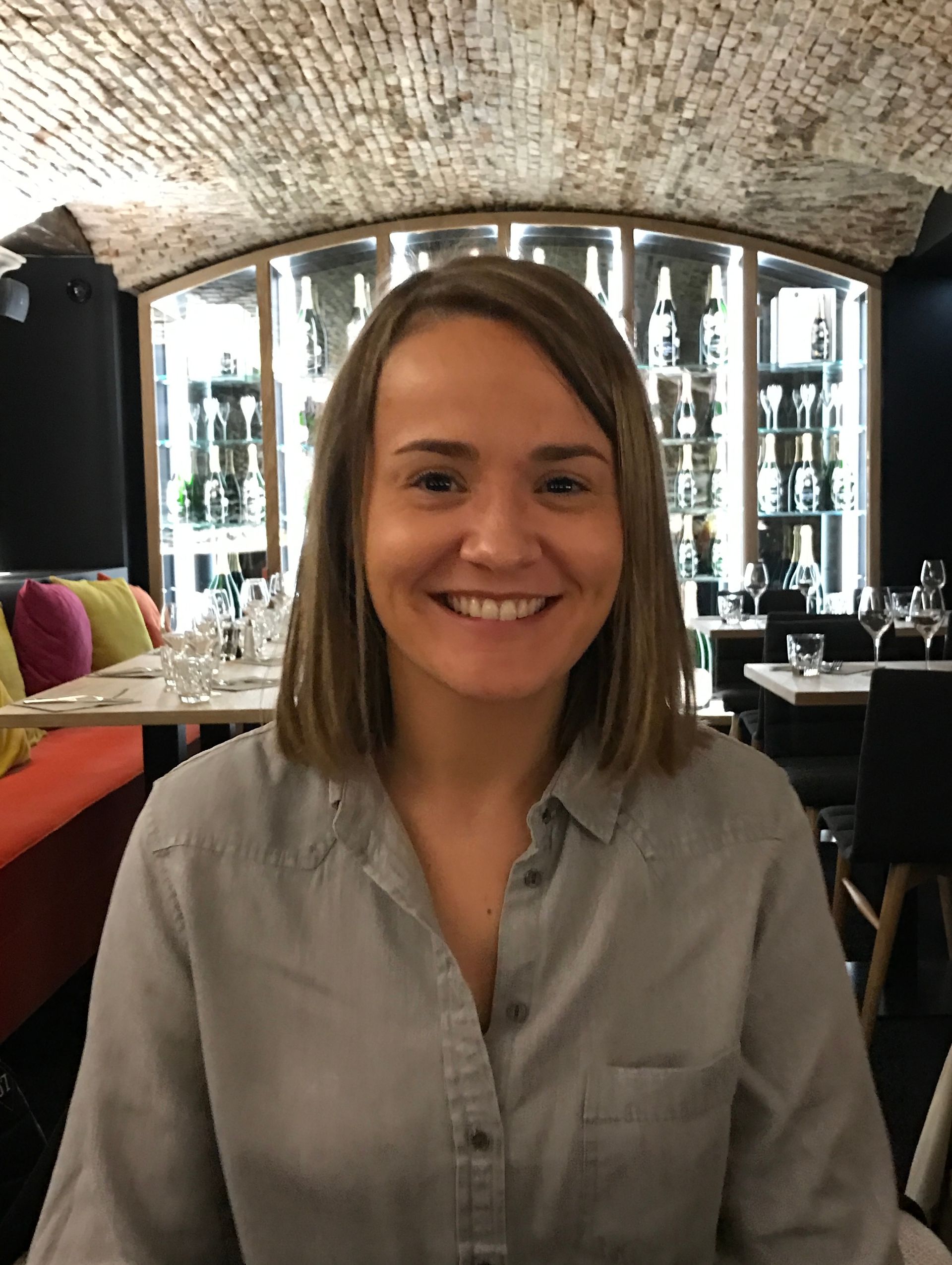 Femme souriante dans un restaurant, vêtue d'une chemise grise, avec un plafond voûté en briques et une cave en arrière-plan.