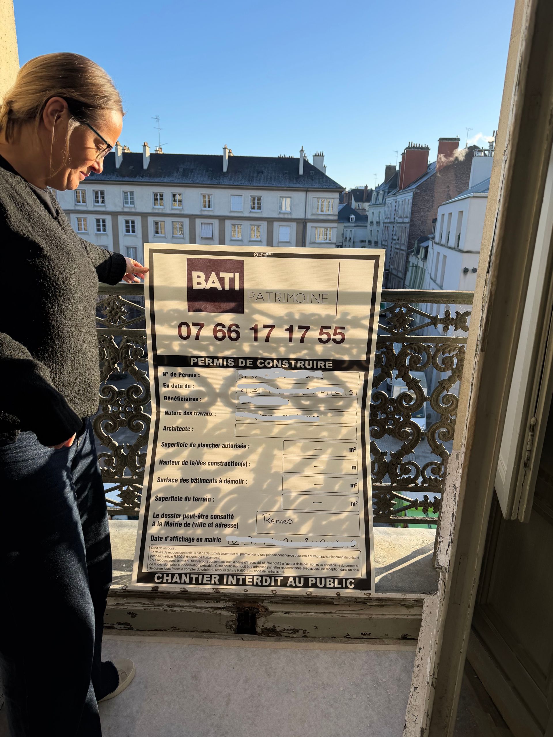 Une femme regarde un panneau de chantier depuis un balcon donnant sur des immeubles.