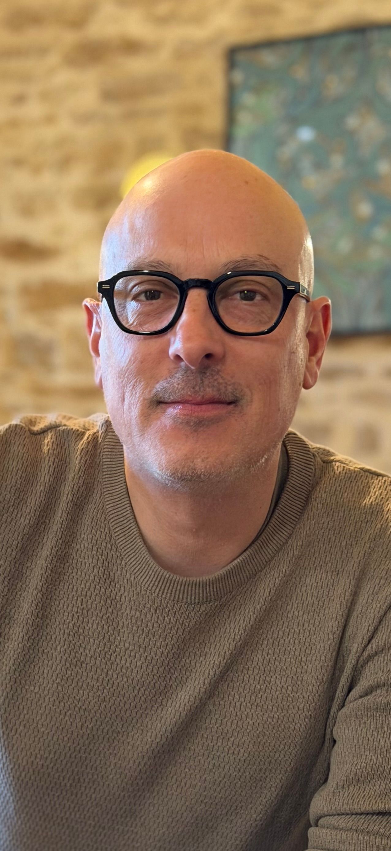 Un homme chauve portant des lunettes et un pull clair se tient devant un mur de pierre.