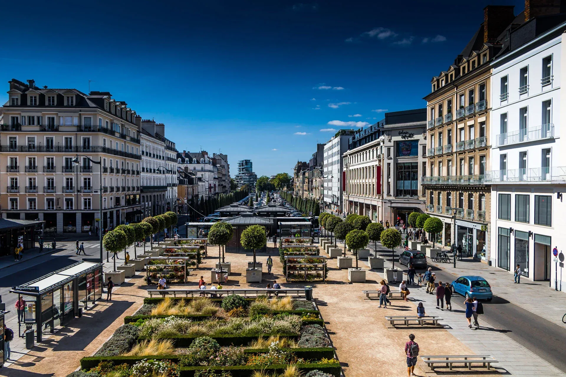 Scène de rue spacieuse avec des bâtiments et une zone piétonne où l'on croise des gens.