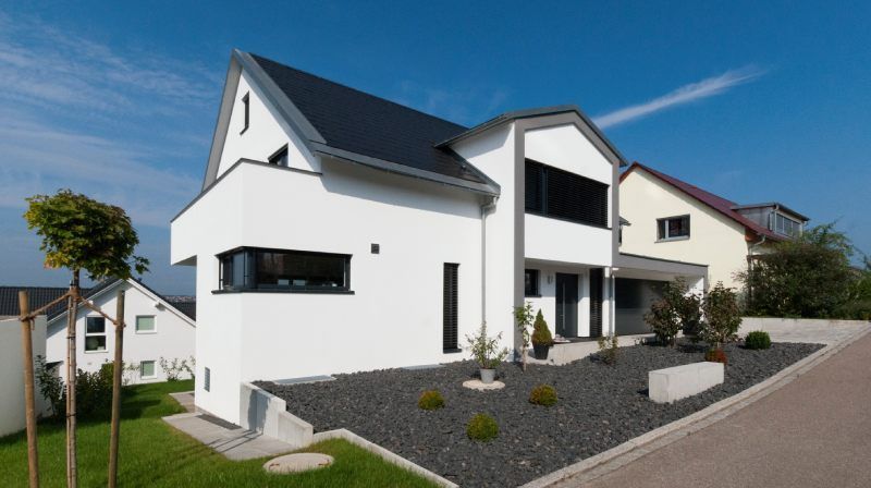 Modernes weißes Haus mit dunklem Dach, schwarzer Kiesfläche und blauem Himmel.