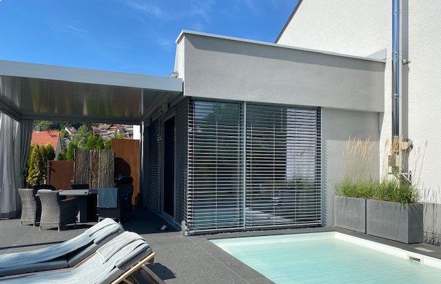Terrasse mit Pool, Liegestuhl und überdachtem Essbereich; moderne Gebäudefassade mit grauen Jalousien und blauem Himmel.