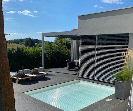 Terrasse mit Pool, modernes Haus, Liegestühle, Pergola und bewölkter Himmel.