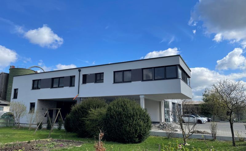 Modernes weißes Gebäude mit dunkel gerahmten Fenstern unter einem blauen Himmel mit flauschigen Wolken.