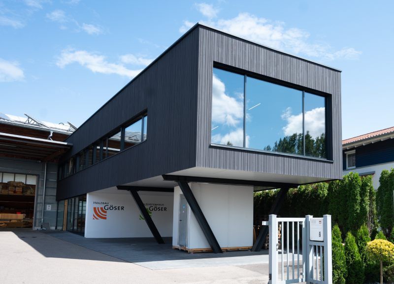 Modernes, dunkelgraues Gebäude mit großen Fenstern, getragen von schrägen Metallträgern. Sonniger Tag, weiße Wand mit Logo.