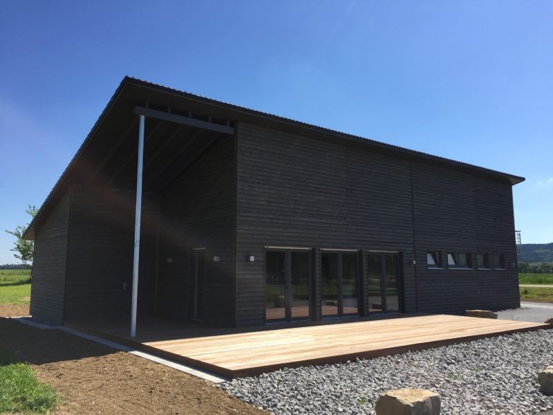 Schwarzes, modernes Gebäude mit Holzterrasse, Glastüren und schrägem Dach an einem sonnigen Tag.