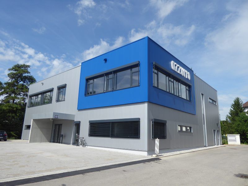 Modernes Gebäude mit blau-grauer Fassade, Logo „izzini“, unter blauem Himmel.