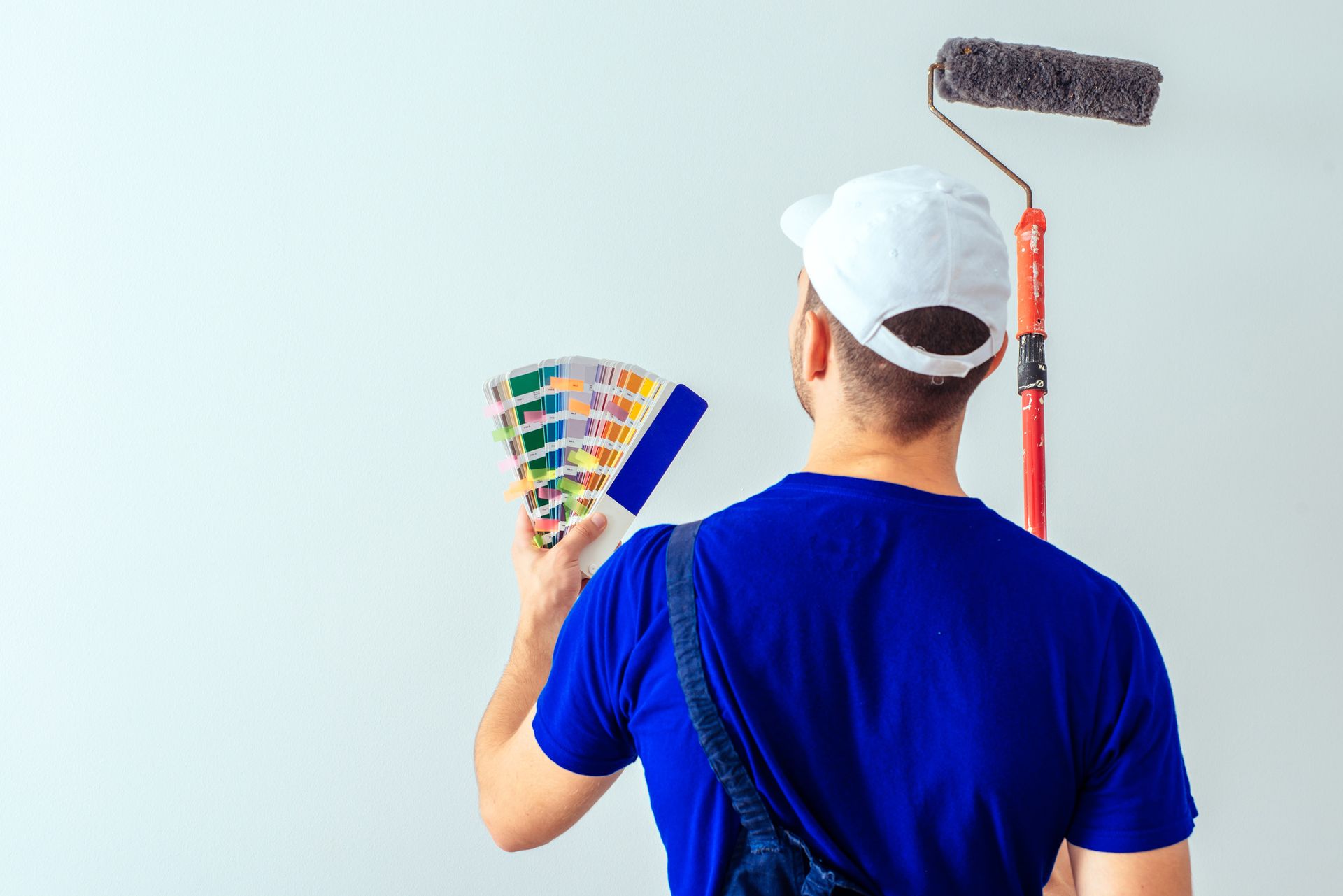 Pintor con ropa azul y gorra blanca, elige un color de pintura de una paleta mientras sostiene un rodillo de pintura.