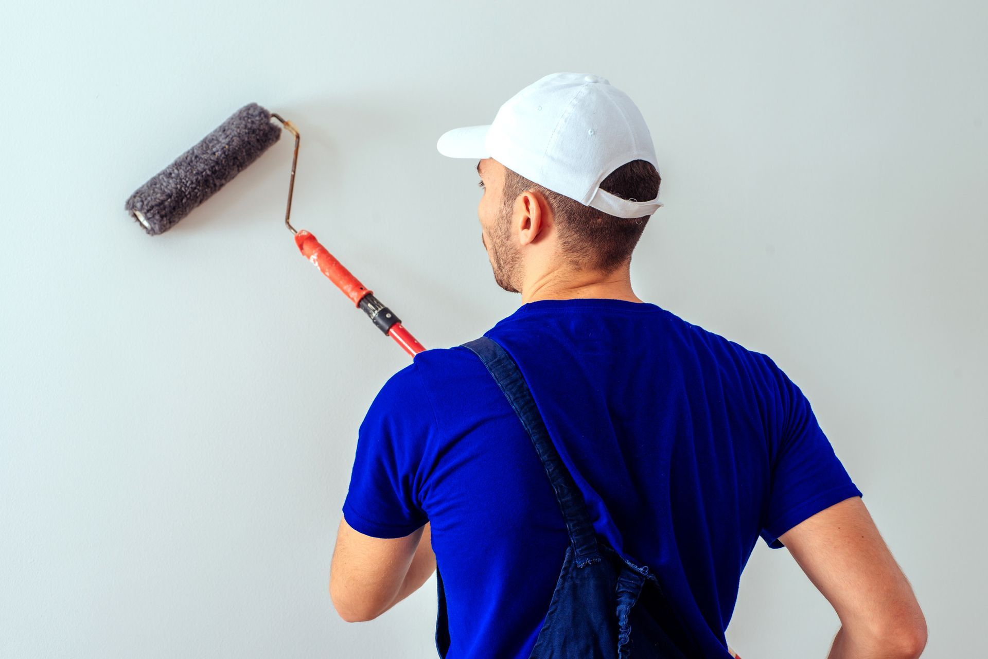 Hombre pintando una pared de blanco con un rodillo, vestido con un mono azul y una gorra blanca.