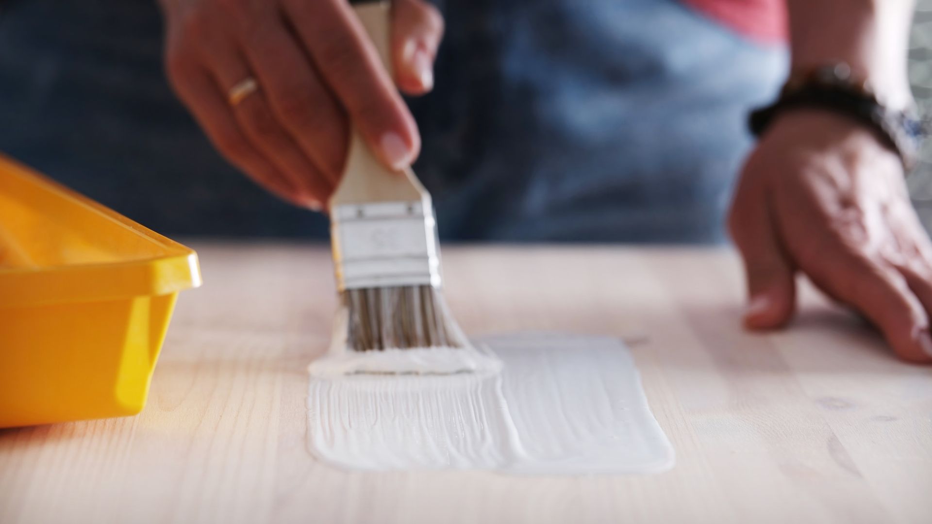 Persona aplicando pintura blanca sobre madera con un pincel; bandeja de pintura amarilla visible.
