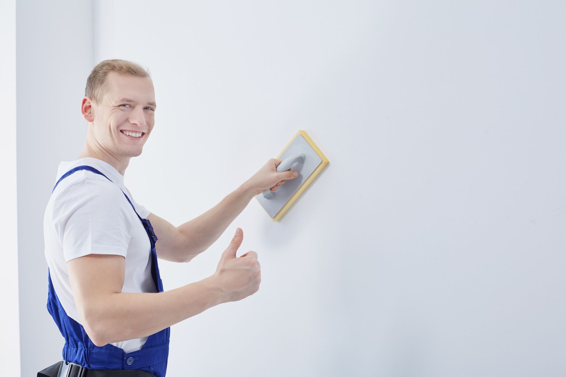 Un hombre con un mono azul alisa una pared blanca con un bloque de lijado y hace un gesto de aprobación con el pulgar.