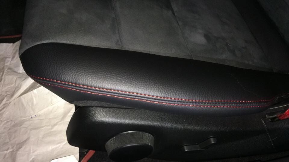 Detalhe de um banco de carro. Lateral em couro preto com costura vermelha, parte superior em camurça cinza e botão preto.