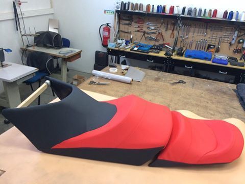 Banco de motocicleta, em couro vermelho e preto, sobre uma bancada em uma oficina com ferramentas e suprimentos.