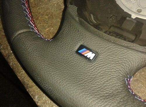 Volante de carro em couro preto com logotipo M e costura tricolor.