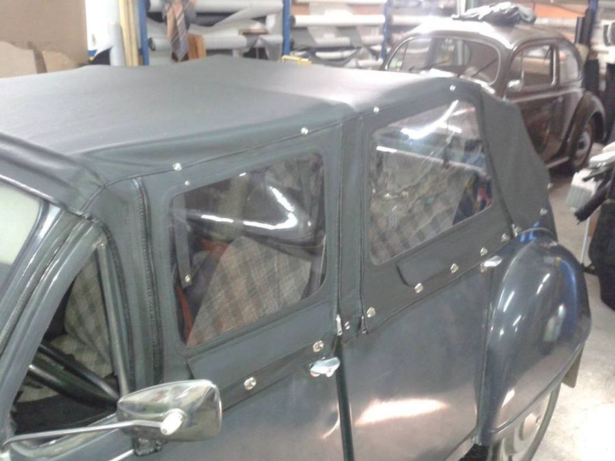 Capota conversível preta com janelas transparentes, carroceria escura e carro antigo ao fundo.