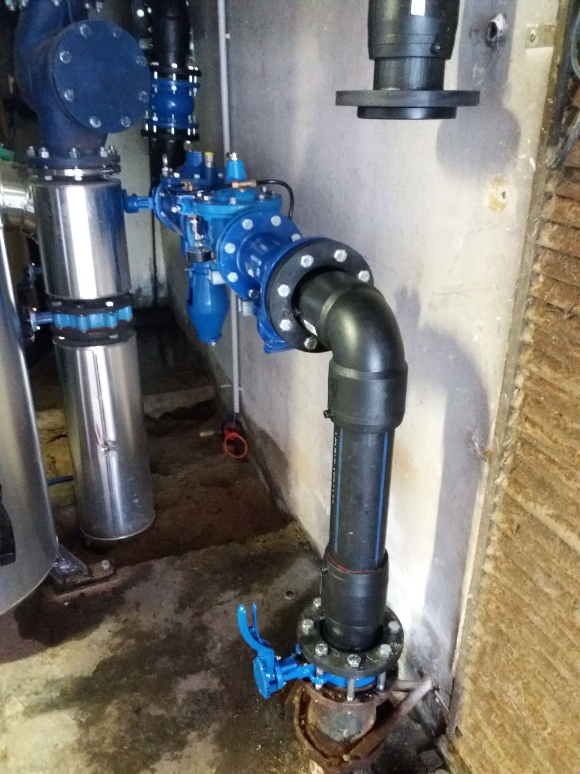 Main gantée ajustant les raccords de plomberie, avec le mur bleu et les tuyaux métalliques visibles.
