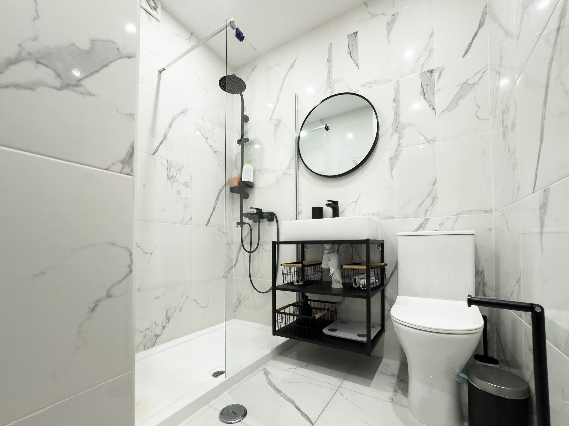 Salle de bains moderne avec murs en marbre blanc, douche à l'italienne, miroir rond, meuble vasque, toilettes et poubelle