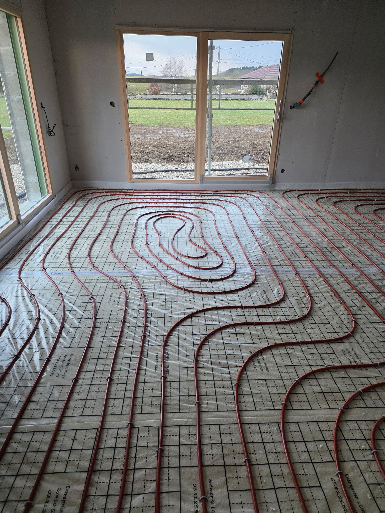 Une pièce avec des tuyaux de chauffage au sol rouges posés sur l'isolant, donnant sur une porte-fenêtre.