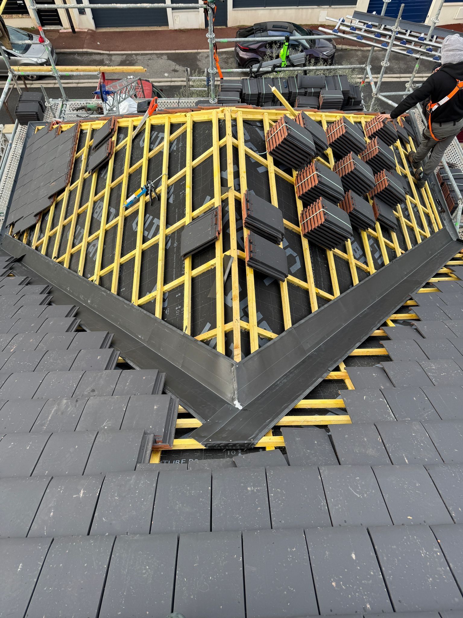 Une personne portant un équipement de sécurité installe des tuiles de toit sombres sur une charpente en bois, sur un toit d'habitation partiellement construit.