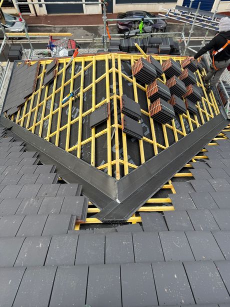 Une personne portant un équipement de sécurité installe des tuiles de toit sombres sur une charpente en bois, sur un toit d'habitation partiellement construit.