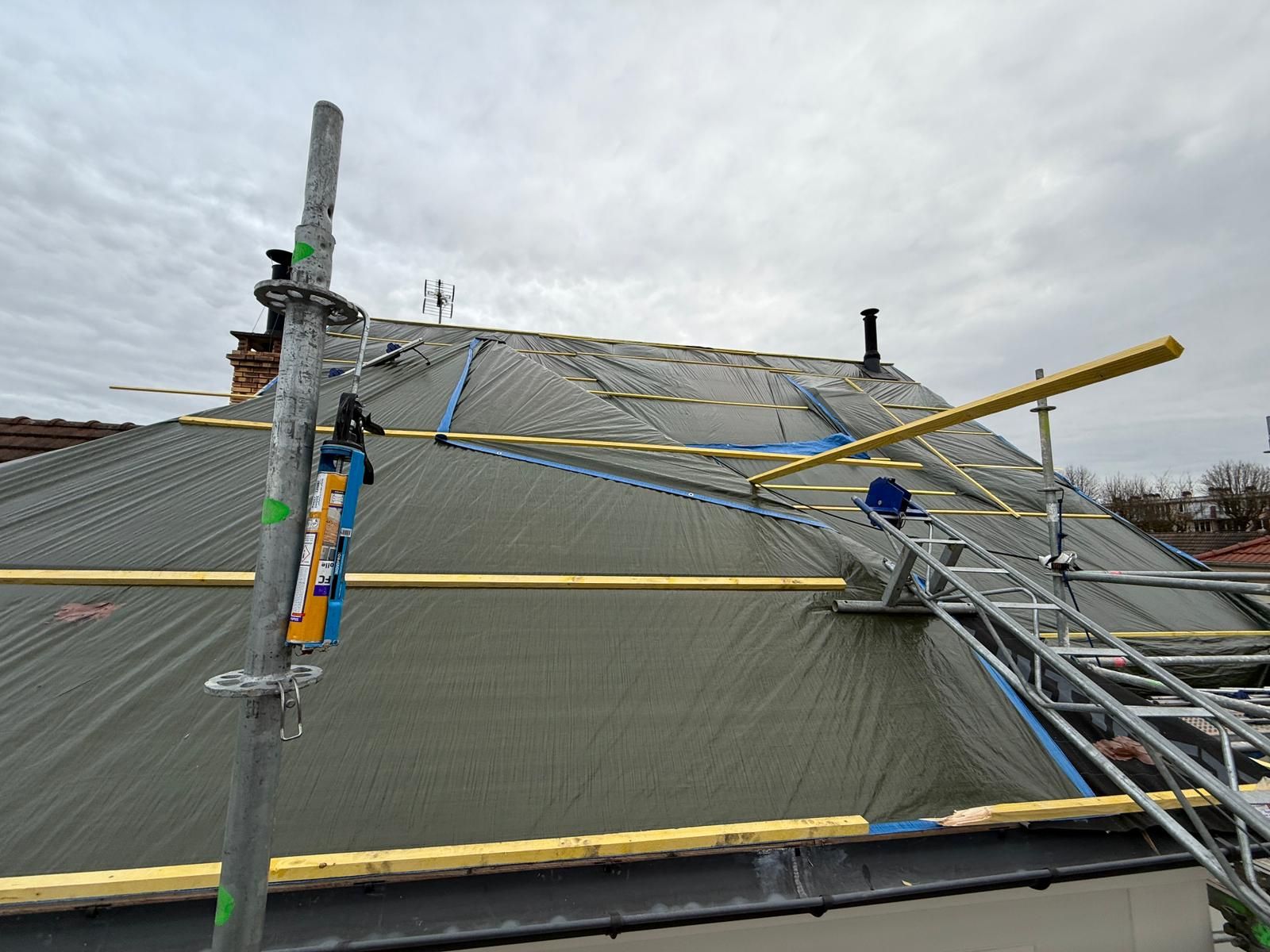 Un toit pentu en construction, recouvert de bâches imperméables grises, de poutres de soutien en bois et d'échafaudages métalliques.