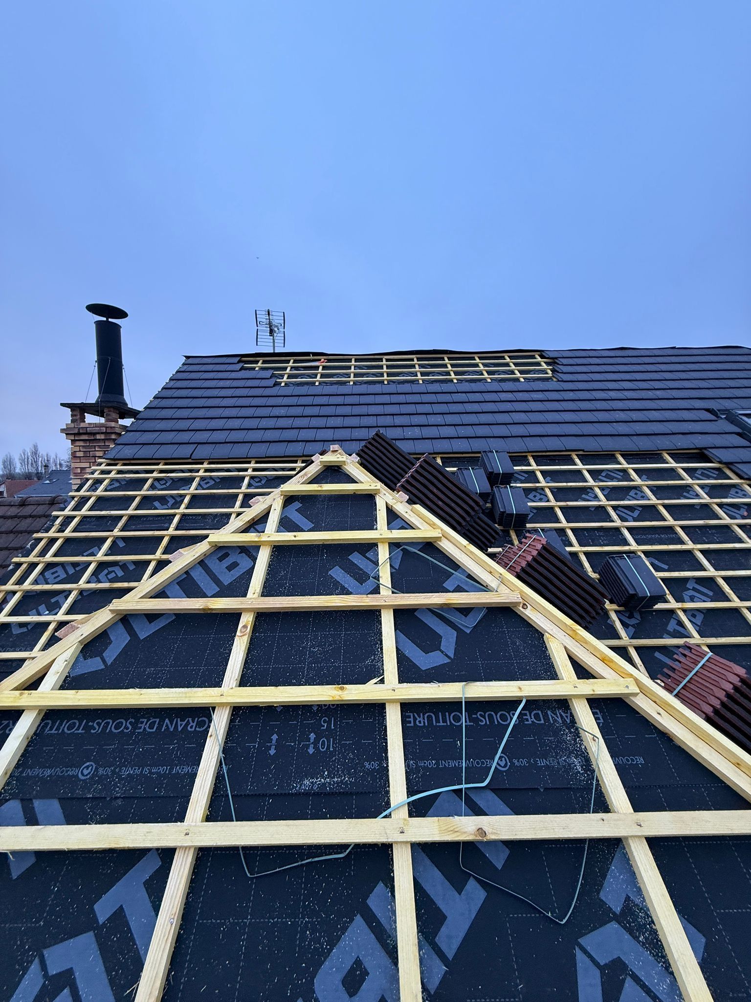 Un toit d'habitation en cours de réparation, montrant des lattes de bois, une sous-couche noire et des piles de tuiles neuves.