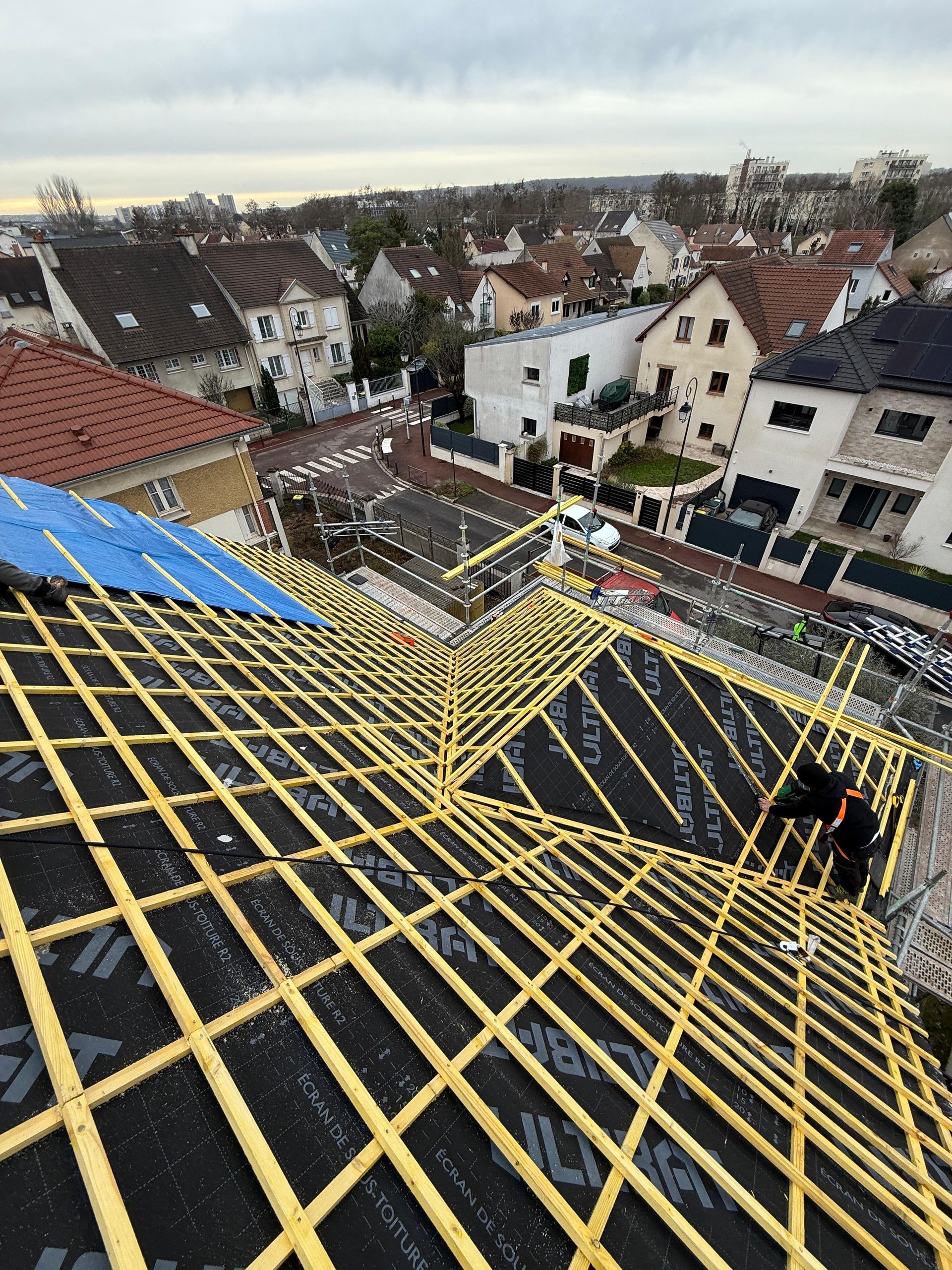 Vue en plongée d'un toit en construction, montrant des lattes de charpente en bois jaune installées sur une couche protectrice noire.
