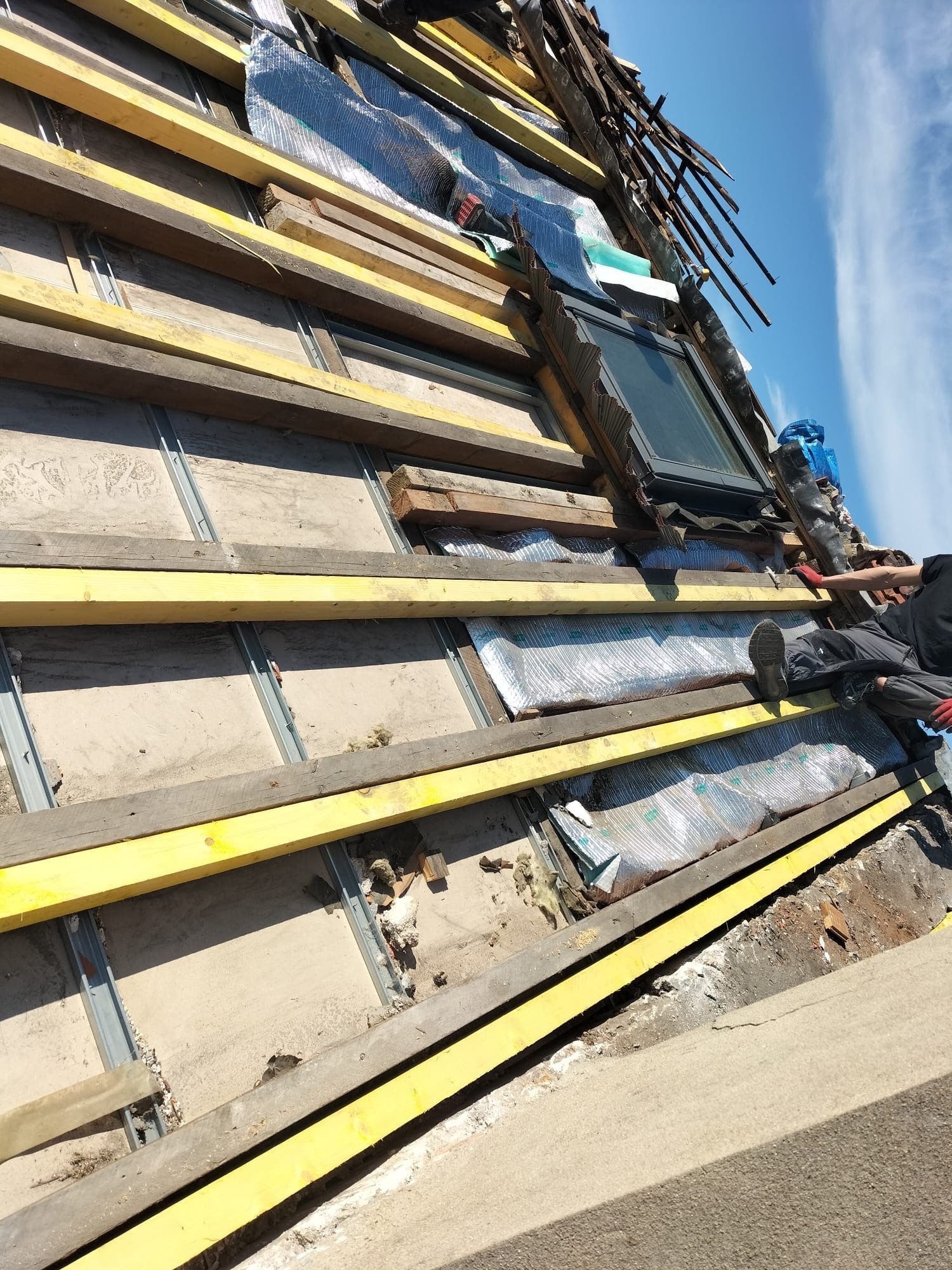 Un ouvrier, juché sur une échelle, installe des liteaux en bois sur une charpente de toit recouverte d'isolant et d'un film plastique protecteur.