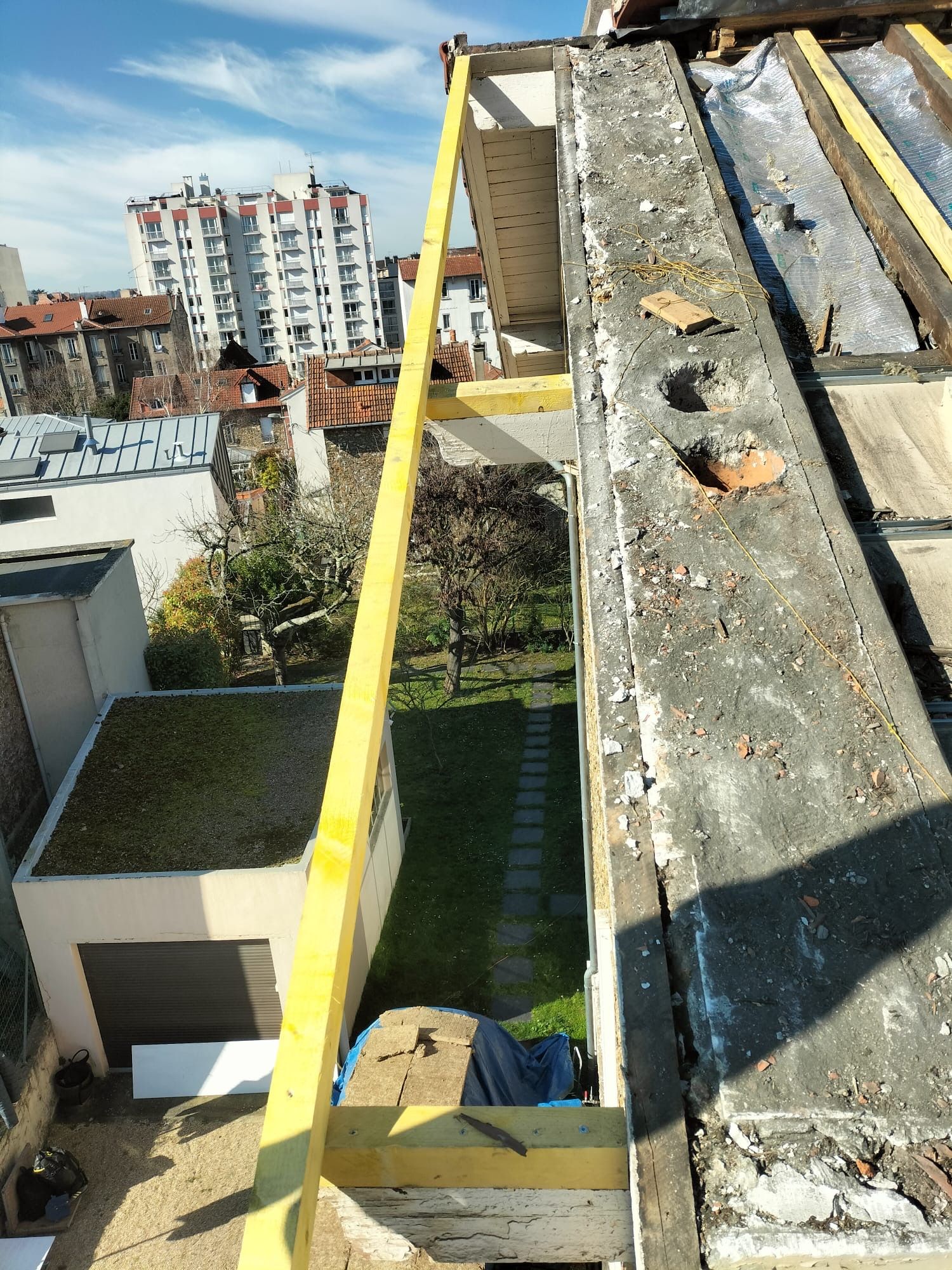 Vue depuis un chantier de construction sur un toit montrant une poutre de sécurité jaune, un bord en pierre endommagé et une cour arrière de banlieue en contrebas.