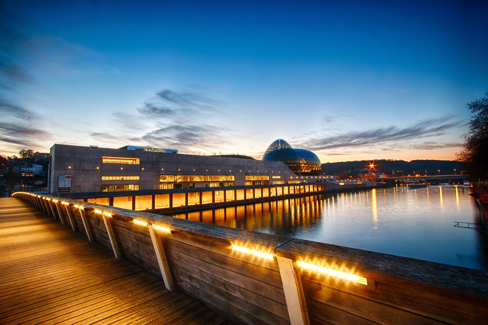 Une passerelle en bois dotée de rampes éclairées mène à un bâtiment moderne en forme de dôme qui se reflète dans l'eau au crépuscule.